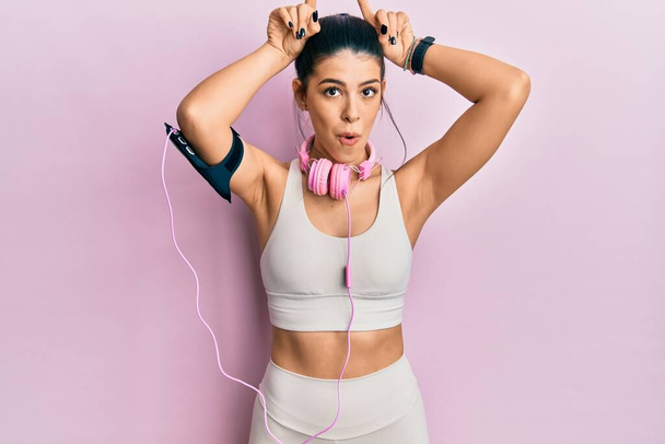 Jeune femme hispanique portant des vêtements de gymnastique et utilisant des écouteurs faisant un geste drôle avec le doigt sur la tête comme cornes de taureau  - Photo, image
