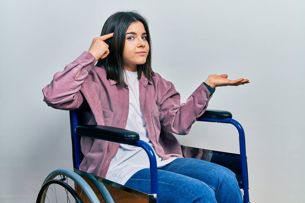 Молодая брюнетка, сидящая на инвалидном кресле, сбита с толку и раздражена открытой ладонью, показывающей пространство для копирования и указывающей пальцем на лоб. think about it.  - Фото, изображение