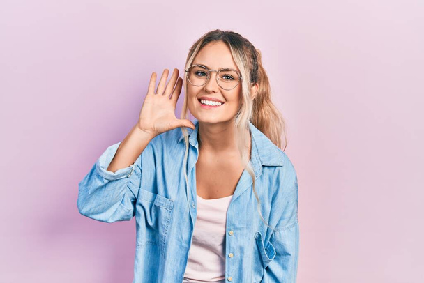 Mooie jonge blonde vrouw met casual kleding en een bril lachend met hand op oor luisterend naar geruchten of roddels. doofheid.  - Foto, afbeelding