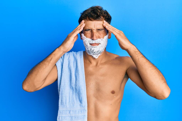 Hombre guapo salvando la barba con espuma afeitada sobre la cara que sufre de dolor de cabeza desesperado y estresado debido al dolor y la migraña. manos en la cabeza.  - Foto, Imagen