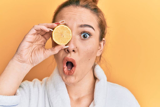 Mladá blondýna žena v županu drží citron přes oko vystrašený a ohromený s otevřenými ústy pro překvapení, nevěřící tvář  - Fotografie, Obrázek
