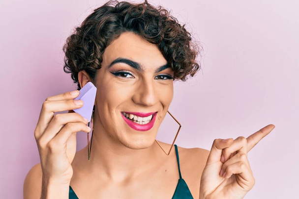 Young man wearing woman make up holding makeup sponge smiling happy pointing with hand and finger to the side  - Foto, immagini