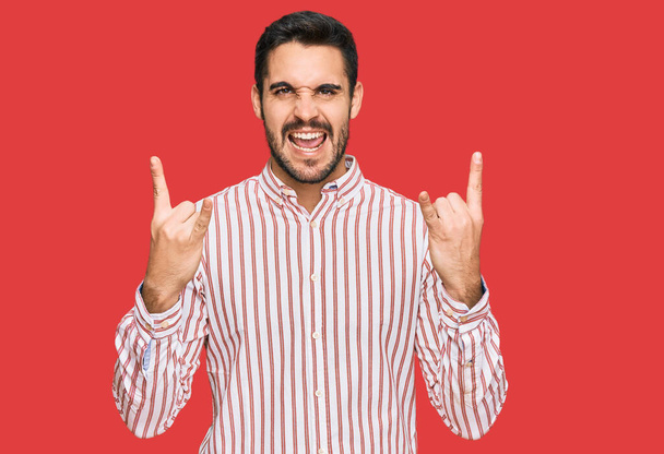 Joven hombre hispano con camisa de negocios gritando con expresión loca haciendo símbolo de roca con las manos arriba. Estrella de la música. concepto pesado.  - Foto, imagen