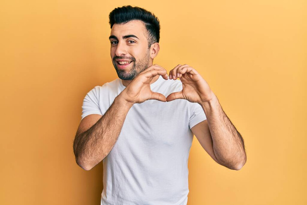 Jeune homme hispanique portant un t-shirt blanc décontracté souriant en amour faisant forme de symbole de coeur avec les mains. concept romantique.  - Photo, image