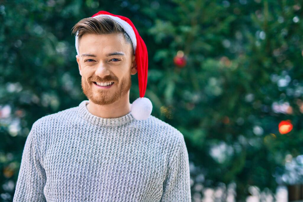 Parkta mutlu gülümseyen genç beyaz adam Noel şapkası takıyor.. - Fotoğraf, Görsel