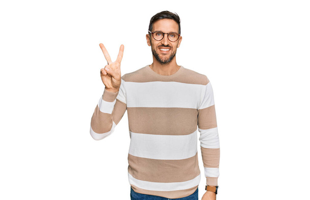 Hombre guapo con barba llevando ropa casual y gafas mostrando y apuntando hacia arriba con los dedos número dos mientras sonríe confiado y feliz.  - Foto, imagen