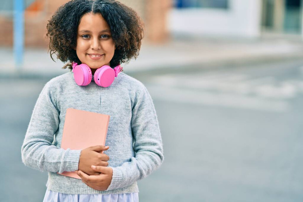 Αξιολάτρευτη ισπανόφωνη μαθήτρια που κρατάει βιβλίο και χρησιμοποιεί ακουστικά στην πόλη. - Φωτογραφία, εικόνα
