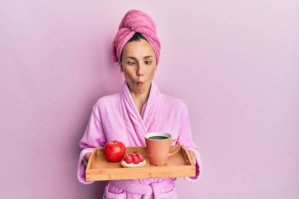 Young blonde woman wearing bathrobe holding tray with breakfast making fish face with mouth and squinting eyes, crazy and comical.  - Photo, Image