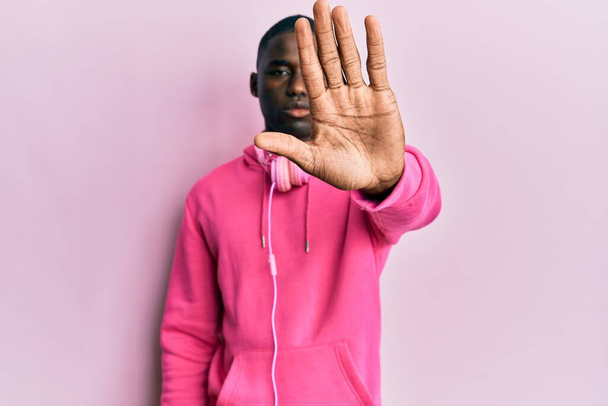 Young african american man wearing gym clothes and using headphones doing stop sing with palm of the hand. warning expression with negative and serious gesture on the face.  - Photo, Image