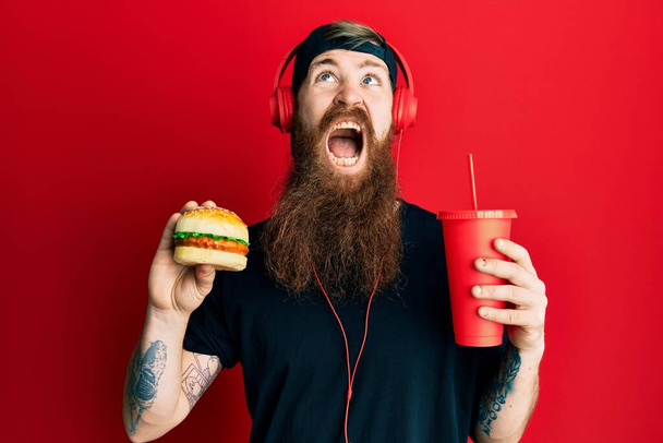 Rudowłosy mężczyzna z długą brodą jedzący smacznego klasycznego hamburgera i pijącego sodę wściekłego i szalonego krzyczącego sfrustrowanego i wściekłego, krzyczącego ze złości patrzącego w górę.  - Zdjęcie, obraz
