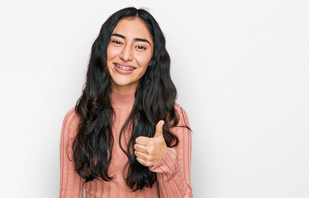 Hispanisches Teenager-Mädchen mit Zahnspange in lässiger Kleidung, das mit der Hand fröhlich Daumen nach oben streckt. Zustimmender Blick in die Kamera zeigt Erfolg.  - Foto, Bild