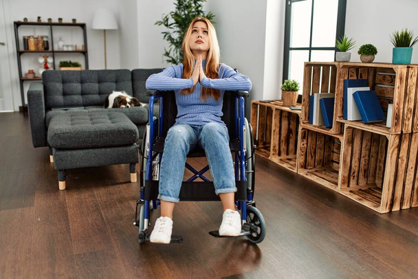 Young beautiful woman sitting on wheelchair at home begging and praying with hands together with hope expression on face very emotional and worried. begging.  - Fotoğraf, Görsel