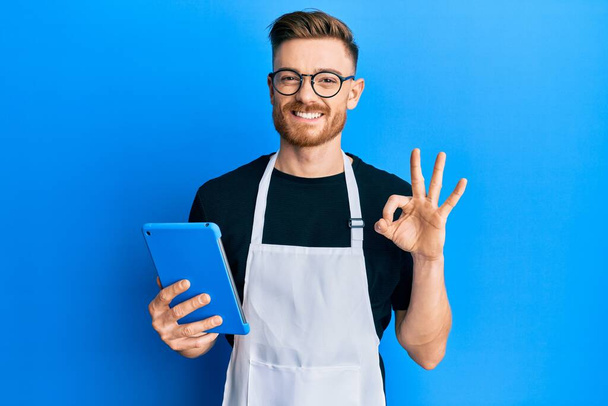 Νεαρός κοκκινομάλλης άνδρας φορώντας επαγγελματική ποδιά χρησιμοποιώντας touchpad κάνει ok υπογράψει με τα δάχτυλα, χαμογελώντας φιλικό gesturing εξαιρετικό σύμβολο  - Φωτογραφία, εικόνα