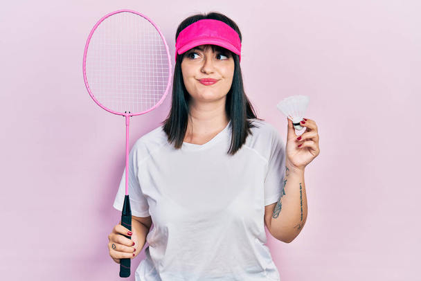 Joven mujer hispana sosteniendo raqueta de bádminton y lanzadera sonriendo mirando hacia un lado y mirando hacia otro pensando.  - Foto, imagen