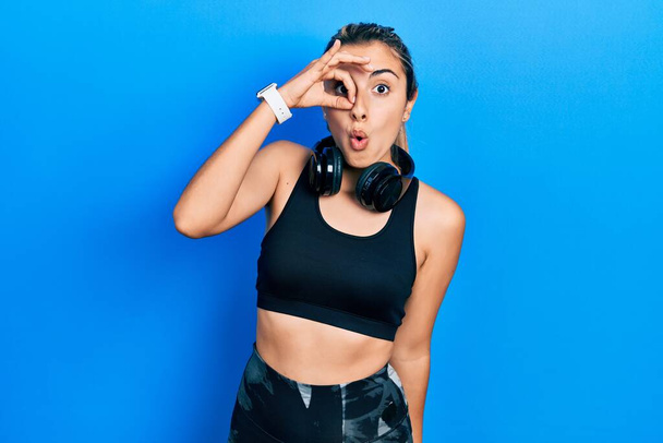Beautiful hispanic woman wearing gym clothes and using headphones doing ok gesture shocked with surprised face, eye looking through fingers. unbelieving expression.  - Photo, Image