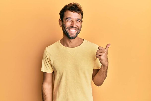 Handsome man with beard wearing casual yellow tshirt over yellow background smiling with happy face looking and pointing to the side with thumb up.  - Photo, Image