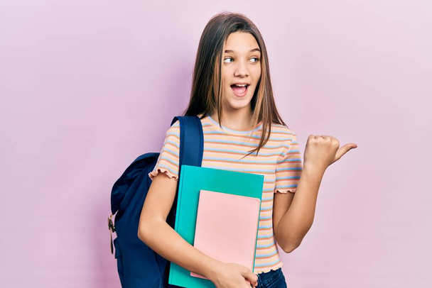 Νεαρή μελαχρινή κοπέλα που κρατάει φοιτητική τσάντα και βιβλία που δείχνουν τον αντίχειρα στο πλάι χαμογελώντας χαρούμενη με ανοιχτό στόμα  - Φωτογραφία, εικόνα