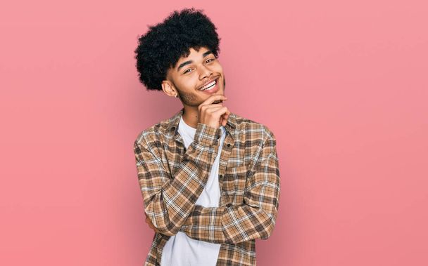 Young african american man with afro hair wearing casual clothes looking confident at the camera with smile with crossed arms and hand raised on chin. thinking positive.  - Фото, изображение
