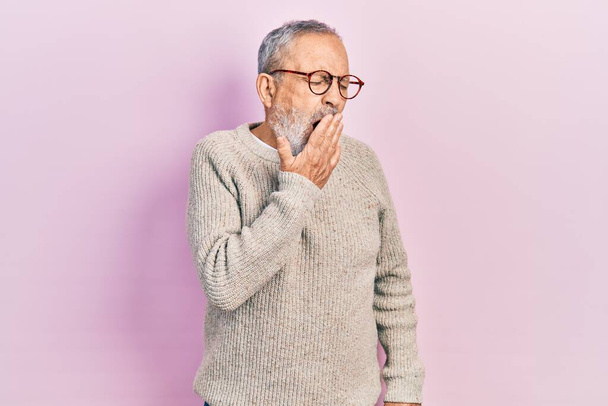 Όμορφος ηλικιωμένος άντρας με γενειάδα που φοράει περιστασιακό πουλόβερ και γυαλιά βαριέται χασμουρητό κουράστηκε να καλύπτει το στόμα με το χέρι. ανησυχία και υπνηλία.  - Φωτογραφία, εικόνα