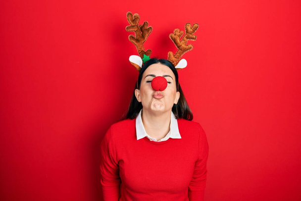 Young hispanic woman wearing deer christmas hat and red nose looking at the camera blowing a kiss on air being lovely and sexy. love expression.  - Photo, Image