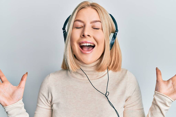 Молодая блондинка слушает музыку, используя наушники, празднующие безумный и сумасшедший успех с поднятыми руками и закрытыми глазами, крича взволнованно. победитель концепция  - Фото, изображение