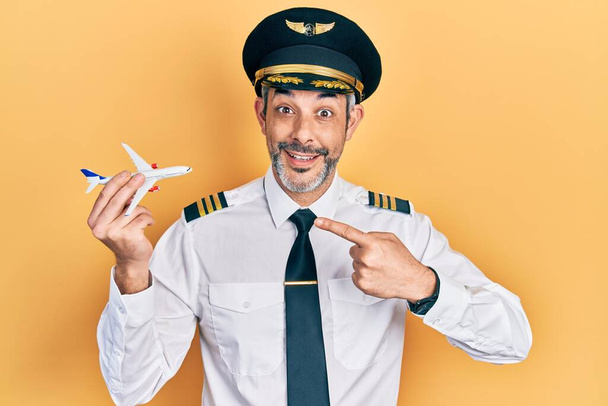 Bello uomo di mezza età con i capelli grigi che indossa uniforme pilota aereo tenendo aereo giocattolo sorridente felice puntando con mano e dito  - Foto, immagini