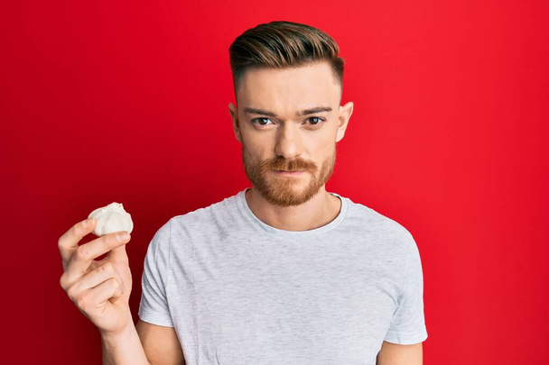 Young redhead man holding fresh garlic thinking attitude and sober expression looking self confident  - Photo, Image
