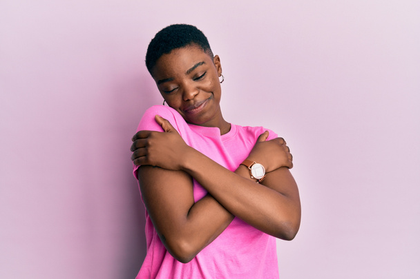 Молода афроамериканська жінка, одягнена в рожеву сорочку, обіймає себе щасливим і позитивним, усміхаючись впевнено. Любов і турбота про себе  - Фото, зображення