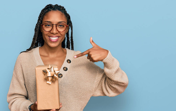 Африканская американка держит в руках подарок, указывая пальцем на себя улыбающийся счастливый и гордый  - Фото, изображение