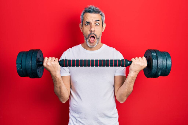Handsome middle age man with grey hair wearing sportswear using dumbbells afraid and shocked with surprise and amazed expression, fear and excited face.  - Photo, Image