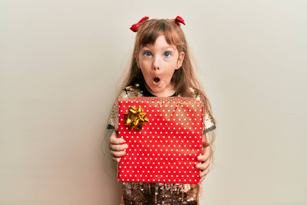 Pequeña niña caucásica sosteniendo regalo rojo asustado y sorprendido con sorpresa y expresión asombrada, miedo y cara excitada.  - Foto, Imagen