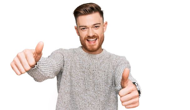 Young redhead man wearing casual winter sweater approving doing positive gesture with hand, thumbs up smiling and happy for success. winner gesture.  - Photo, Image