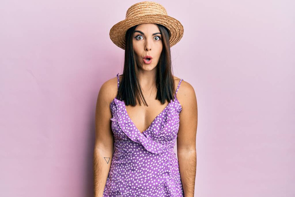 Young brunette woman wearing summer dress and hat afraid and shocked with surprise expression, fear and excited face.  - Photo, Image