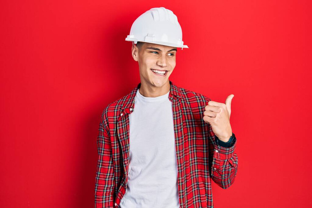 Молодой латиноамериканец в архитектурной каске улыбается со счастливым лицом и указывает на сторону с большим пальцем вверх.  - Фото, изображение
