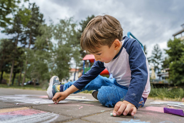 Kleiner kaukasischer Junge im Alter von vier Jahren sitzt im Park auf dem Betonpflaster und zeichnet mit Kreide ein pädagogisches Konzept - Foto, Bild