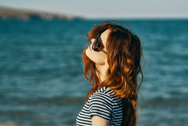 zrzavá žena v slunečních brýlích na pláži v blízkosti modrého moře a hor v pozadí - Fotografie, Obrázek