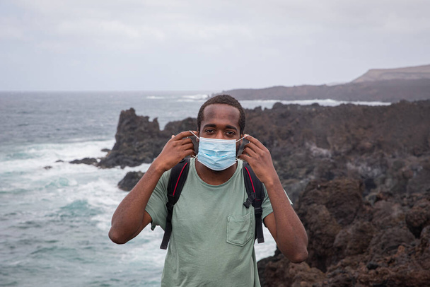 Junger afrikanischer Junge setzt sich auf Lanzarote eine OP-Maske auf und legt seine Hände in die Nähe des Meeres - Foto, Bild