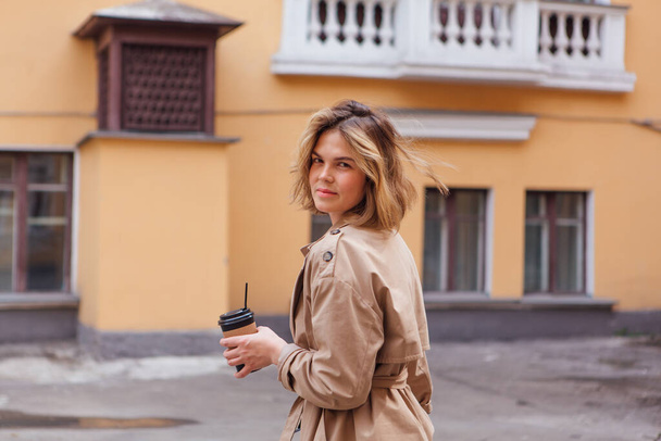 Young smiling millennial woman with wild hair dressed in an autumn coat walking with a cup of coffee to go near the old building. - Foto, afbeelding