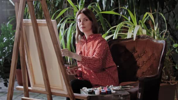 Kunstenaar in een atelier - jonge mooie vrouw tekent een schilderij met kleuren - kijkend in de camera - Video