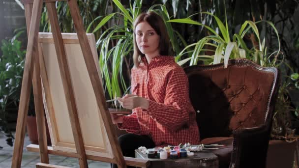 Kunstenaar in een atelier - jonge mooie vrouw tekent een schilderij - kijkend in de camera - Video