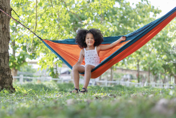 kindertijd en ontspannen concept - weinig afrikaanse amerikaanse krullend haar meisje liggend en genietenon op hangmat kleur oranje onder grote boom. - Foto, afbeelding