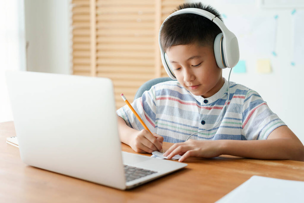 Aasialainen pieni poika videopuhelu verkossa Internetin kautta tutor tietokoneen kannettavan kuulokkeella. Aasialainen tekee läksyjä. Verkko-oppimisen käsite kotona  - Valokuva, kuva