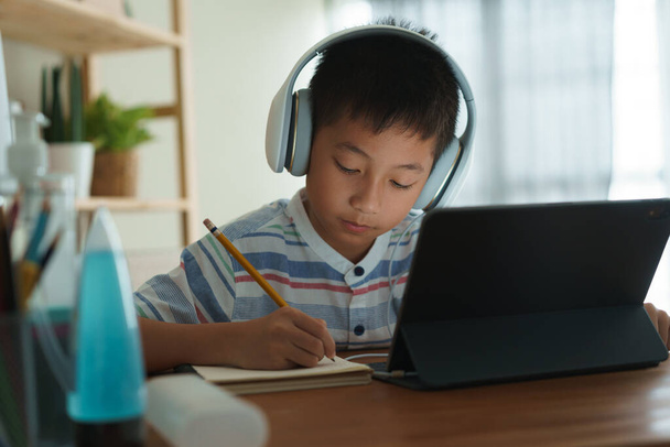 Ασιάτης αγόρι μαθαίνει online μέσω Διαδικτύου με έναν δάσκαλο σε ένα tablet ψηφιακό με ακουστικά, παιδί της Ασίας μελετάει ενώ κάθεται στο σαλόνι στο σπίτι. Έννοια της online μάθησης στο σπίτι  - Φωτογραφία, εικόνα