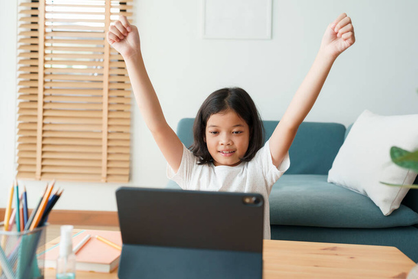 Happy Asian little girl raise hand is learning online with a tablet while sitting in living room in morning, Concept distance learning at home - Φωτογραφία, εικόνα