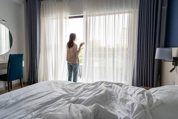 Young asian woman holding cup of coffee and opening curtains in the morning and looking through the window at home - Foto, immagini