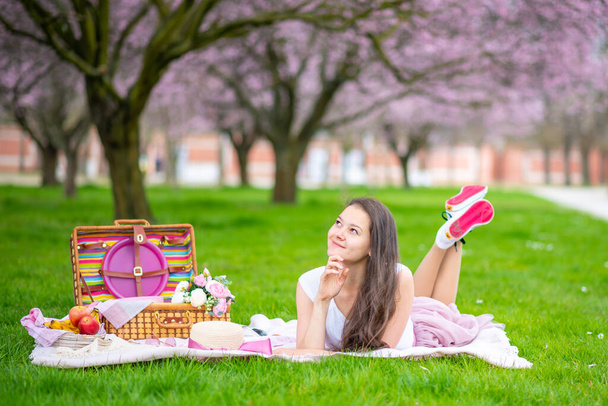 Hermosa mujer en sombrero de paja bajo cerezos florecientes en lugar de picnik. Chica usando una falda larga rosa en el jardín de primavera - Foto, Imagen
