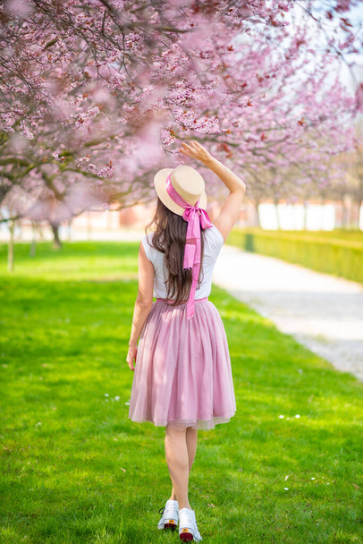 Gyönyörű nő szalmakalapban sétál egy nyári kertben virágzó cseresznyefákkal. Rózsaszín szoknyás lány - Fotó, kép