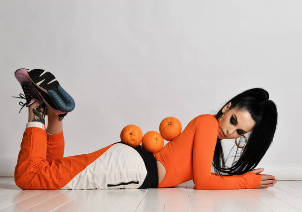 Young slim long-haired brunette woman in orange sportswear and sneakers is lying on floor with fresh oranges on back - Фото, изображение