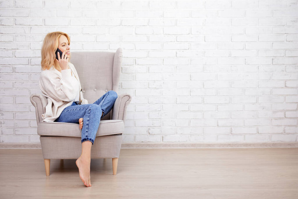 young beautiful woman sitting on armchair and talking by smart phone - copy space over white brick wall background - Fotoğraf, Görsel