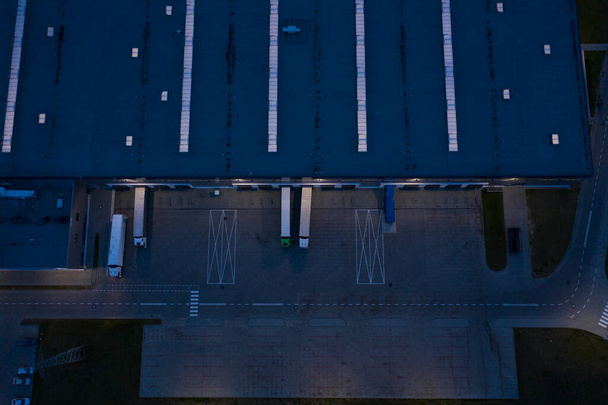 Vista aérea de almacén de mercancías en la noche. Centro logístico en zona industrial de ciudad desde arriba. Vista aérea de camiones cargando en el centro logístico. Vista desde el dron. - Foto, Imagen